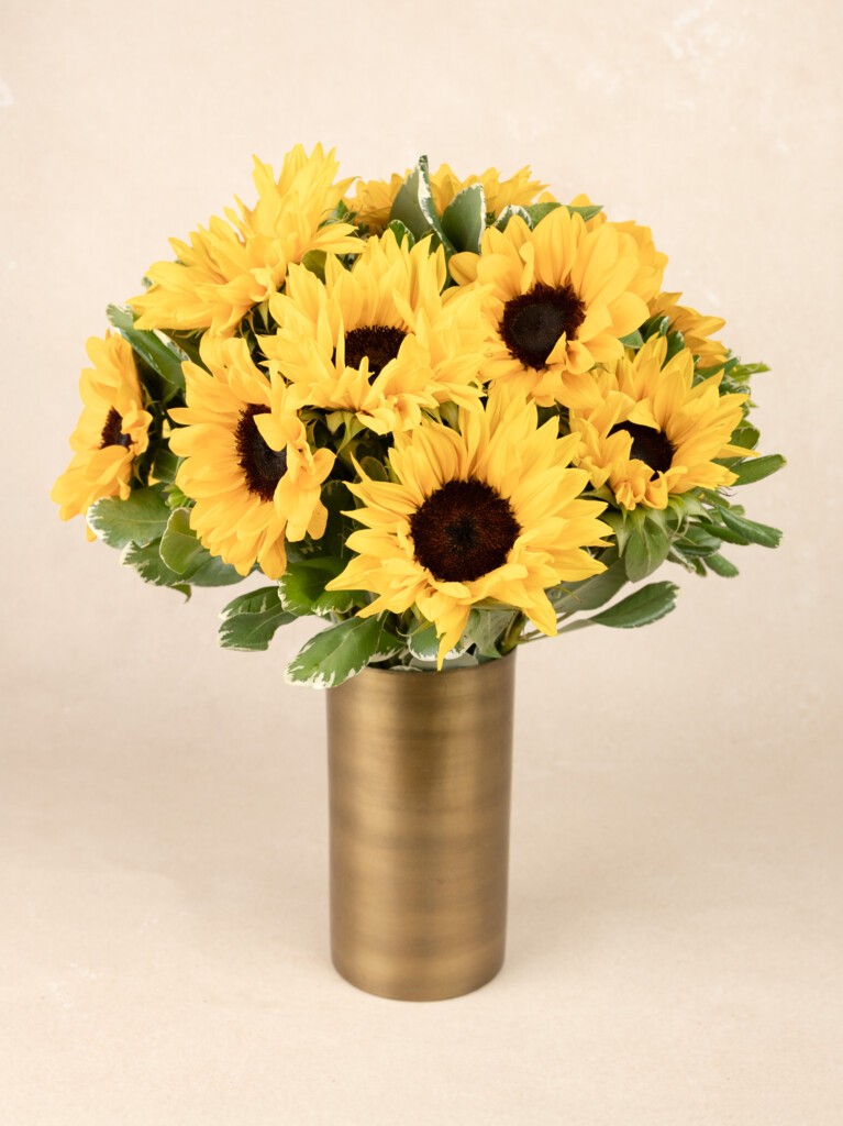 A full, round bouquet of bright yellow sunflowers with dark centers and lush green foliage, arranged in a tall, brushed gold cylindrical vase.