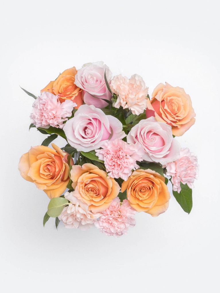 Top-down view of a tightly arranged bouquet featuring apricot-toned roses, pale pink roses, and delicate pink carnations, revealing the layered textures and warm pastel palette in full bloom.