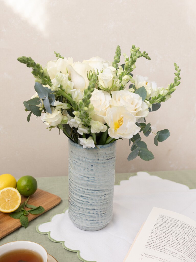 Paulette bouquet with 21 white blooms styled on a table, featuring roses, snapdragons, and eucalyptus. Ideal for stylish home decor or relaxed celebrations.