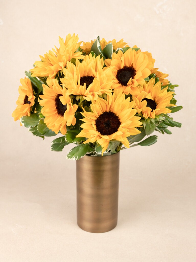 A full, round bouquet of bright yellow sunflowers with dark centers and lush green foliage, arranged in a tall, brushed gold cylindrical vase.
