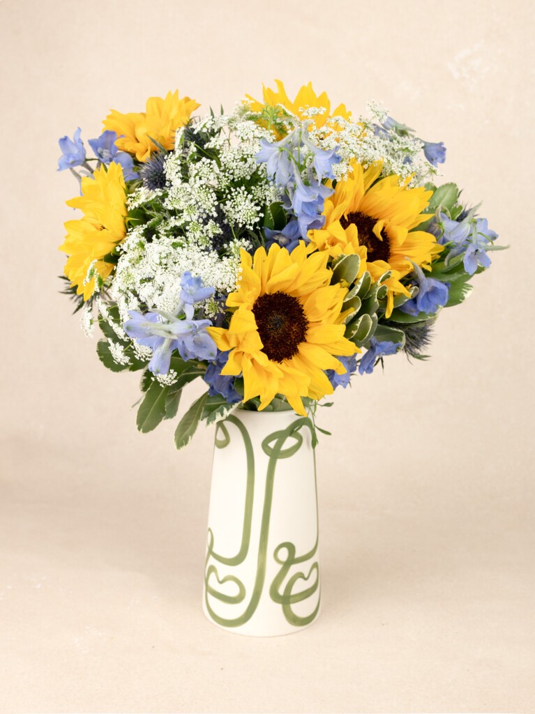 A cheerful sunflower arrangement styled in a white artistic vase, bursting with blues, whites, and sunny yellows for a radiant, countryside-inspired display.
