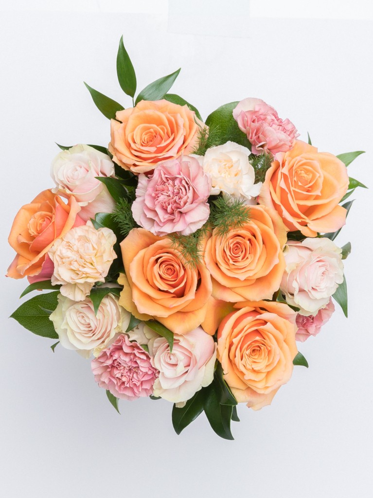 A compact, rounded bouquet photographed from above, highlighting the symmetrical placement of orange, pink, and ivory blooms with pops of greenery nestled between the flowers.