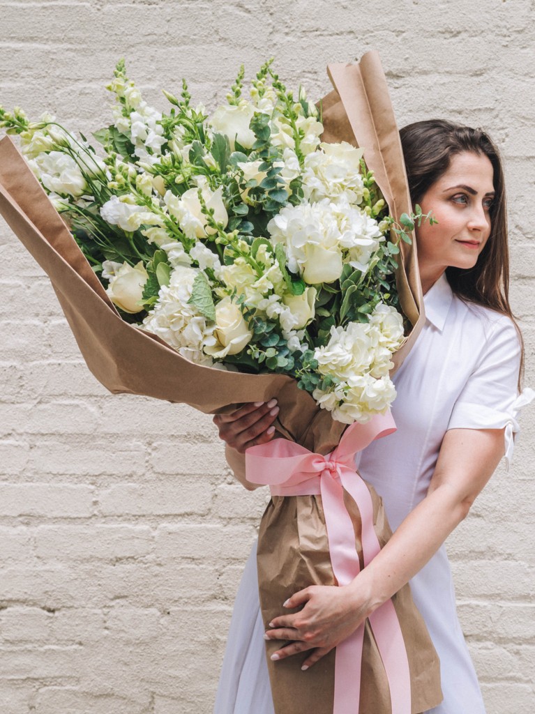 Close side view of a woman holding an oversized bouquet of white and cream blooms wrapped in kraft paper, highlighting the soft textures, sculptural fullness, and striking, armful size of the arrangement.