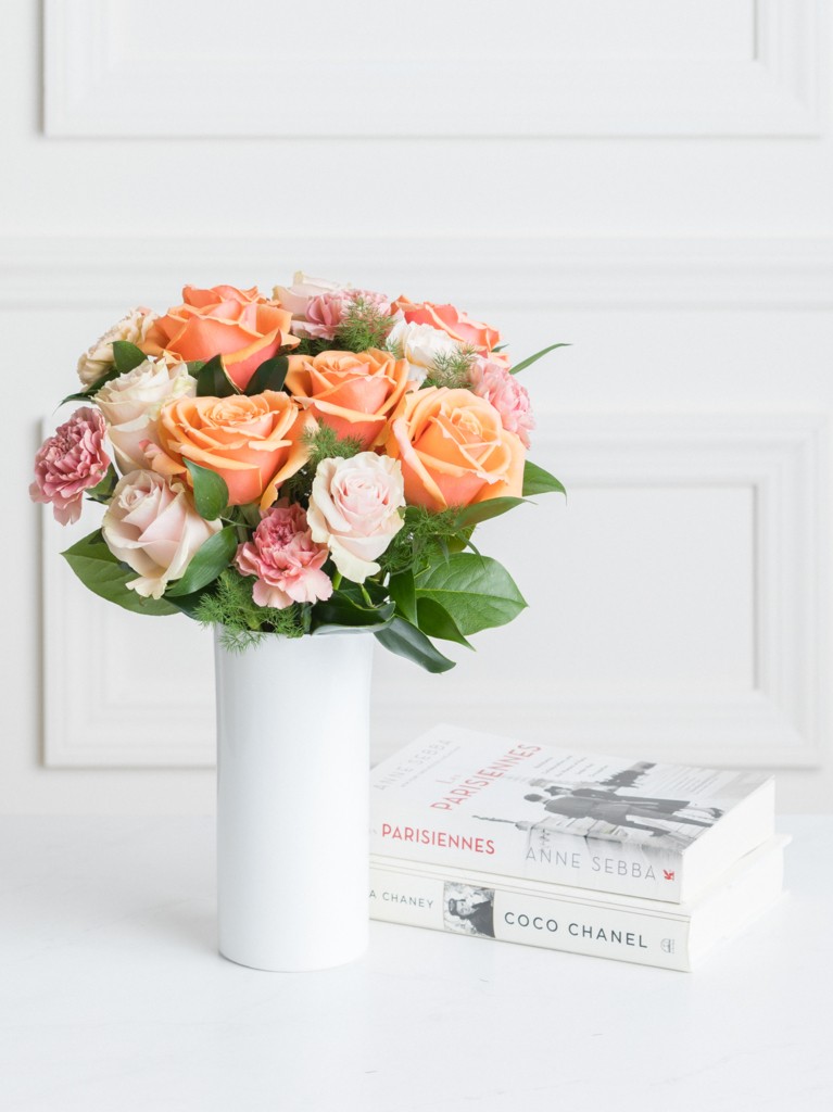 A vibrant arrangement of coral, blush, and cream roses paired with ruffled carnations, presented in a tall white ceramic vase beside two chic fashion books for a soft, elegant display.