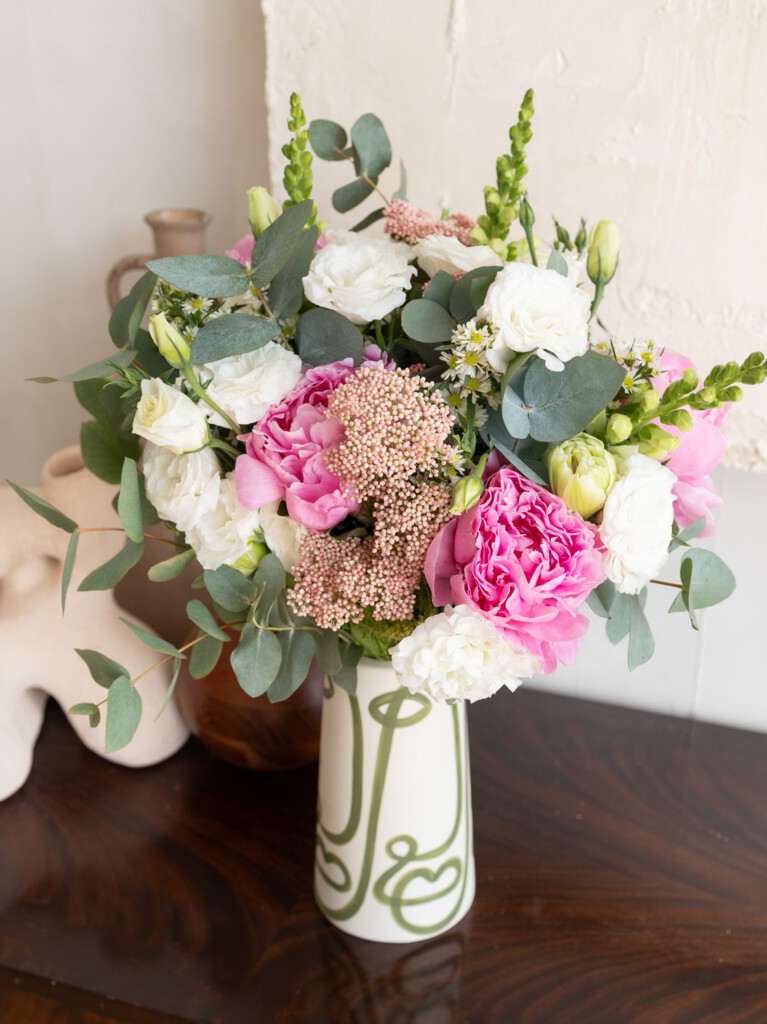 A mixed bouquet displayed in a designer white ceramic vase with green swirl patterns, featuring pink peonies, white lisianthus, pale snapdragons, eucalyptus leaves, and clusters of pale pink rice flowers.