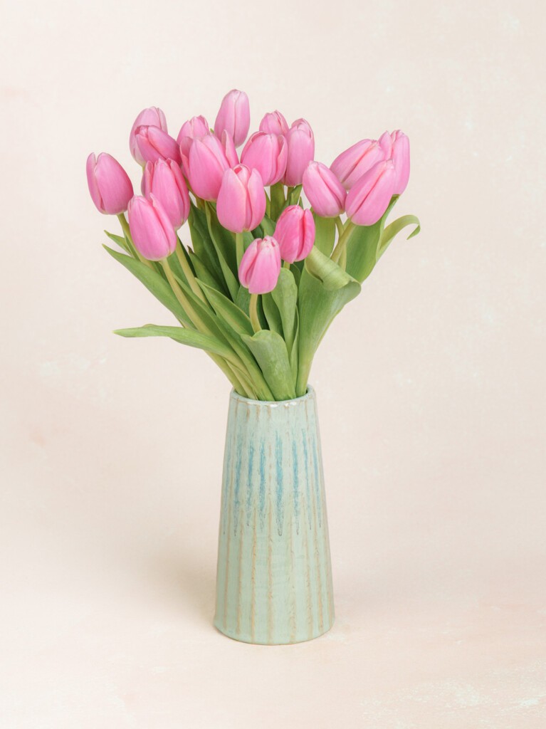 A modern white vase holds 20 vibrant pink tulips with upward stems and vivid green leaves, artfully spilling out in a loose, elegant fan.