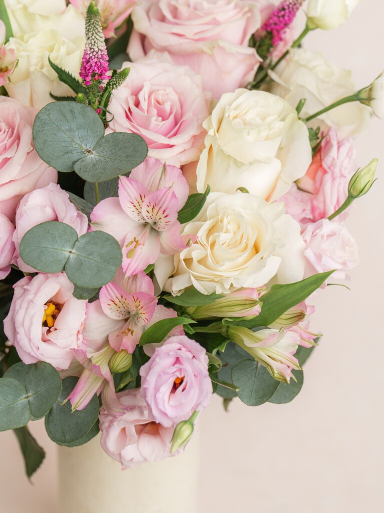 A pink and white floral bouquet in a tall white vase, paired with a spa kit featuring a rustic candle, bath bomb, and skincare essentials, all arranged on a linen produce bag.