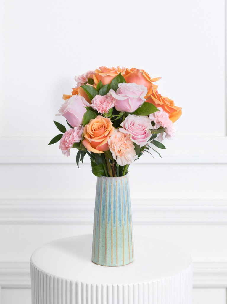 A vertical ceramic vase with soft turquoise and tan striping holds a vibrant bouquet of peach roses, pale pink roses, and blush carnations, arranged with gentle greenery for a balanced, romantic display.
