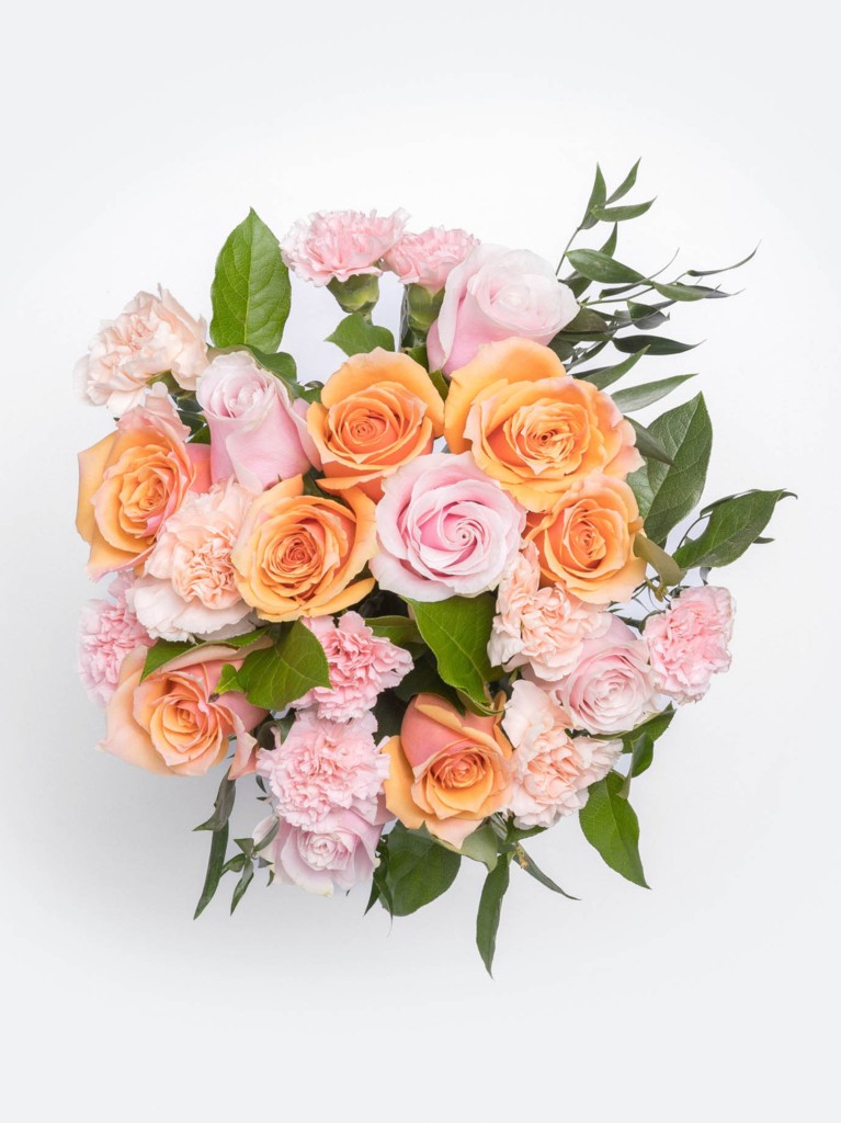 A bouquet viewed from above reveals a harmonious color wheel of apricot, blush, and peach blooms tightly clustered together, interspersed with ruffled carnations and framed by glossy green leaves.