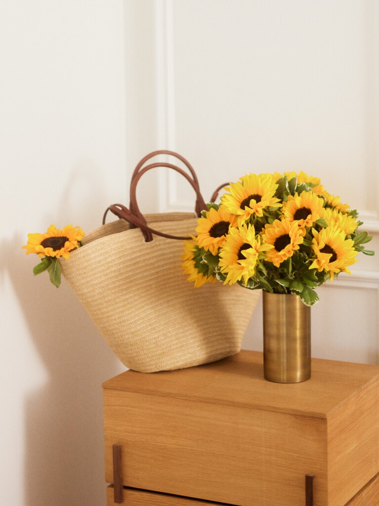 A cheerful bouquet of sunflowers with green foliage in a tall gold vase placed on a wooden cabinet, beside a woven straw tote bag holding two loose sunflower stems.
