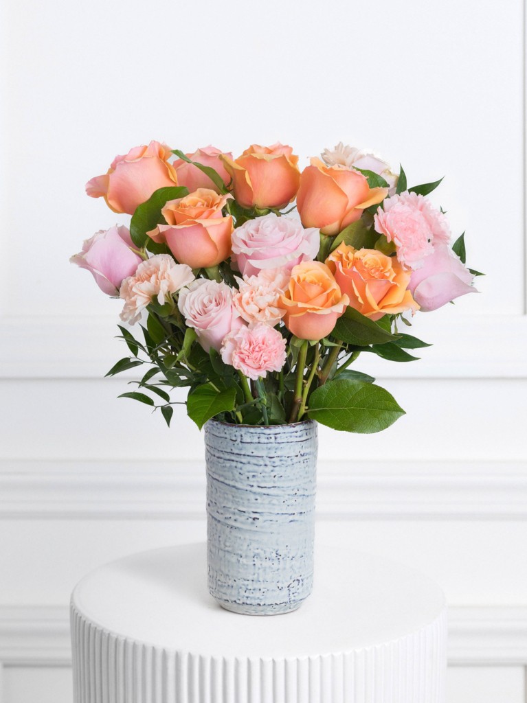 A bright, cheerful bouquet arranged in a textured blue ceramic vase, featuring a lively mix of apricot roses, blush pink roses, pale pink carnations, and soft greenery fanning gently outward in a rounded silhouette.