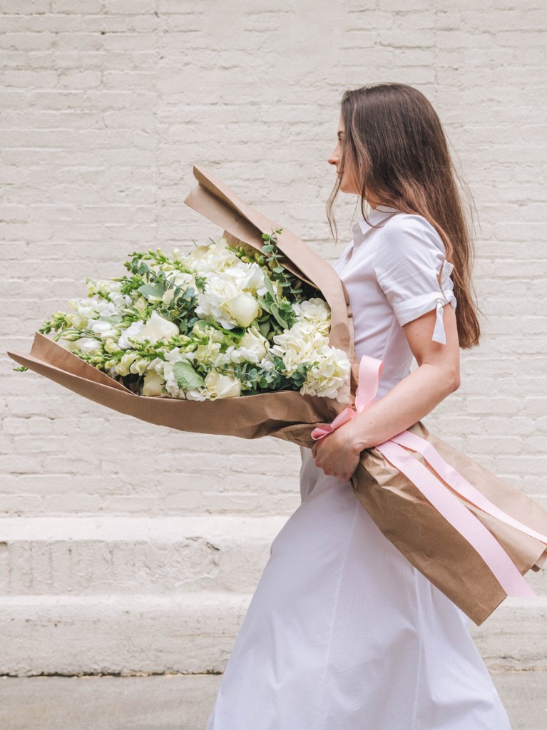 Side view of a woman holding the oversized Sabine bouquet wrapped in kraft paper and ribbon, with cascading white and green seasonal blooms nearly covering her torso to emphasize the bouquet’s dramatic, showstopper scale.